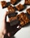 A hand is holding a piece of a dessert bar with layers of rich, dark chocolate and a golden, caramelized nut topping. In the background, several more identical pieces are placed on a surface.