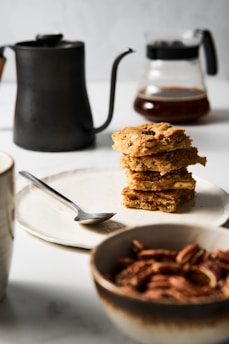 A stack of square, golden-brown cookies sits on a beige plate accompanied by a silver spoon. In the foreground, a bowl is filled with pecans, while in the background, there is a black metal teapot and a glass carafe containing coffee. The setting is minimalist with a neutral-colored background.