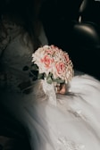 A bride dressed in a white lace wedding gown is holding a bouquet of pink and white roses. The image has a soft, warm lighting that highlights the details of the floral arrangement and the intricate patterns on the bridal dress.