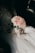 A bride dressed in a white lace wedding gown is holding a bouquet of pink and white roses. The image has a soft, warm lighting that highlights the details of the floral arrangement and the intricate patterns on the bridal dress.