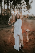 A person dressed in a white gown stands beside a tan horse in a wooded area during sunset. Sunlight filters through the trees, creating a warm ambiance. The scene exudes a sense of tranquility and connection with nature.