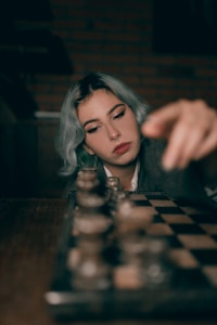 A person with light blue hair is focused on a chessboard, seemingly in the middle of a game. They are pointing towards one of the chess pieces, appearing deep in thought. The background is dimly lit with brick wall details.