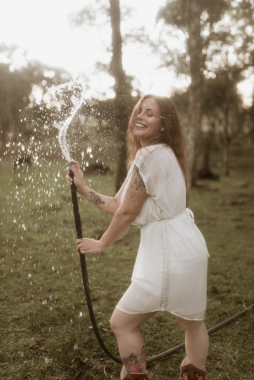 A joyful person with long hair stands outdoors, holding a hose that sprays water, creating a playful and refreshing scene. The person is wearing a white dress and red boots, standing on green grass with trees in the blurred background, giving a serene and natural atmosphere.