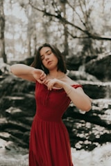 A woman stands in a serene forest setting, wearing a flowing red dress and appearing to be engaged in a moment of introspection or mindfulness with eyes closed. The background features blurred greenery and sunlight filtering through the trees, creating a peaceful ambiance.
