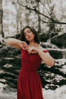 A woman stands in a serene forest setting, wearing a flowing red dress and appearing to be engaged in a moment of introspection or mindfulness with eyes closed. The background features blurred greenery and sunlight filtering through the trees, creating a peaceful ambiance.