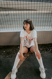 A young person with short dark hair is sitting on the ground with their back against a wire fence. They are wearing a white t-shirt with a graphic print, black shorts, white knee-high socks with red stripes, and white sneakers. The background includes an asphalt surface and a concrete barrier.