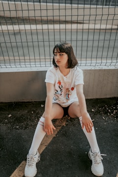 A young person with short dark hair is sitting on the ground with their back against a wire fence. They are wearing a white t-shirt with a graphic print, black shorts, white knee-high socks with red stripes, and white sneakers. The background includes an asphalt surface and a concrete barrier.