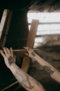 Two tattooed arms reach out towards a wooden structure in an indoor setting with soft lighting, creating a mysterious and artistic atmosphere. The background appears to be blurred, emphasizing the focus on the hands and the tattoos.