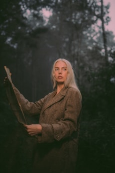 A person with long, light hair stands in a forest holding an open newspaper. The setting appears to be misty and dim, creating a mysterious atmosphere.