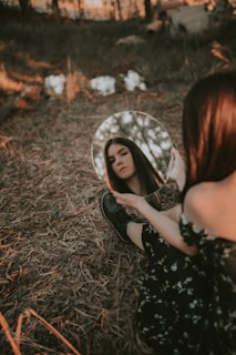 A woman sits on a forest floor covered with dry leaves, holding a round mirror that reflects her face. The surroundings are dimly lit with soft, warm lighting filtering through trees, creating a serene and introspective atmosphere.