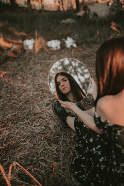 A woman sits on a forest floor covered with dry leaves, holding a round mirror that reflects her face. The surroundings are dimly lit with soft, warm lighting filtering through trees, creating a serene and introspective atmosphere.