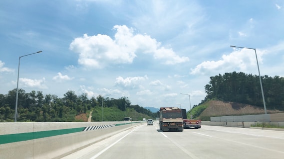 A highway stretches forward under a bright blue sky with scattered fluffy clouds. Trees and shrubbery line the sides of the road, while several vehicles, including a large truck, travel on it. Streetlights are positioned on either side of the road.