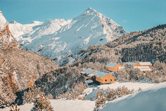 A collection of yellow cabins is nestled among snow-covered hills and a dense forest. The scene is set against a backdrop of towering, snow-capped mountains under a bright blue sky.