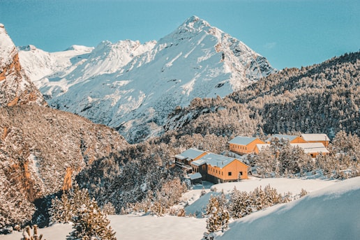 A collection of yellow cabins is nestled among snow-covered hills and a dense forest. The scene is set against a backdrop of towering, snow-capped mountains under a bright blue sky.