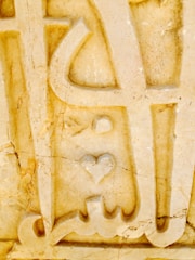 An intricate stone carving featuring abstract shapes and a prominent heart symbol. The surface is textured with visible cracks and variations in color, adding a rustic and weathered appearance.