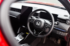 The interior of a car showcasing a steering wheel with the Honda logo prominently displayed. The dashboard is modern with digital screens and various control buttons. The surrounding area includes a gear shift and other car controls, suggesting a high-tech, contemporary vehicle interior.