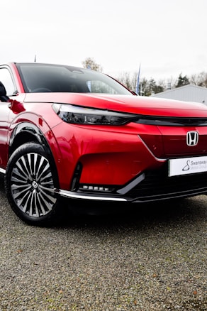 A sleek, modern red car with distinctive black accents and stylish wheels, parked on a gravel surface. The car's design is angular and contemporary, featuring sharp lines and a glossy finish. In the background, there are trees and a building with a light gray color.