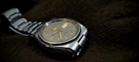 A silver wristwatch with a metal strap and a round, gray dial resting on a dark textured fabric. The watch face features golden hour markers and a day-date window, exuding a classic and precise design aesthetic.