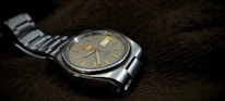 A silver wristwatch with a metal strap and a round, gray dial resting on a dark textured fabric. The watch face features golden hour markers and a day-date window, exuding a classic and precise design aesthetic.