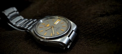 A silver wristwatch with a metal strap and a round, gray dial resting on a dark textured fabric. The watch face features golden hour markers and a day-date window, exuding a classic and precise design aesthetic.