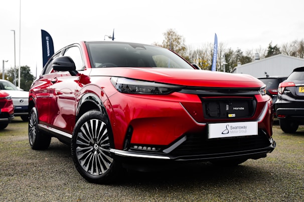 A shiny red SUV with a sleek design is parked outdoors on a gravel surface. The vehicle has modern features including stylish rims and a black trim. In the background, there are other cars and some trees, along with a blue sky and a white building.