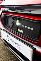 The image features a close-up of an electric car's charging port, with the Honda logo visible. The car is predominantly red with black and white accents. A sign with the text 'Swansway Motor Group' is prominently displayed below the charging area.