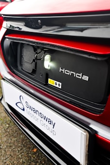 The image features a close-up of an electric car's charging port, with the Honda logo visible. The car is predominantly red with black and white accents. A sign with the text 'Swansway Motor Group' is prominently displayed below the charging area.