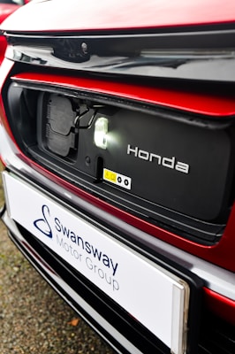 The image features a close-up of an electric car's charging port, with the Honda logo visible. The car is predominantly red with black and white accents. A sign with the text 'Swansway Motor Group' is prominently displayed below the charging area.