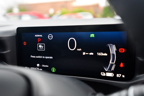 A close-up view of a car's digital dashboard displaying various indicators such as speed, gear position, and mileage. The central panel shows a speed of 0 mph with the gear set to Park. Additional information includes battery charge at 87%, a remaining range of 163 miles, and a prompt to press a switch to operate. The display is primarily black with white, red, and green icons and text.