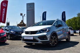 A silver Honda car is prominently displayed in an outdoor dealership lot, surrounded by other vehicles. Large banners and signs with the Honda logo are visible, with a clear blue sky in the background. The lot surface appears to be gravel or asphalt, and there is visible greenery on the side.