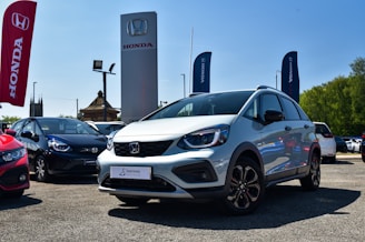A silver Honda car is prominently displayed in an outdoor dealership lot, surrounded by other vehicles. Large banners and signs with the Honda logo are visible, with a clear blue sky in the background. The lot surface appears to be gravel or asphalt, and there is visible greenery on the side.