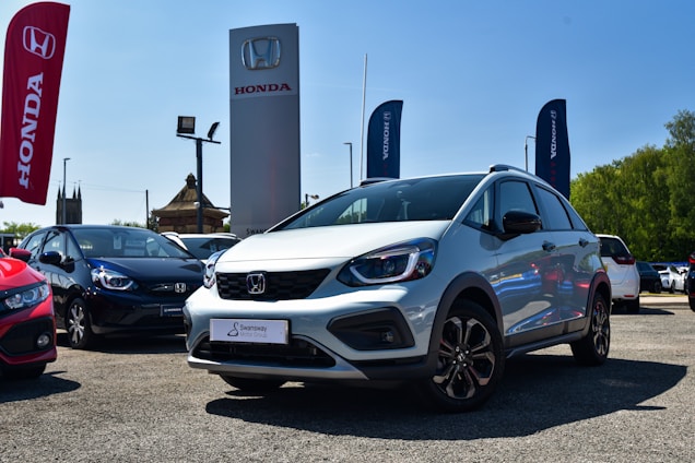 A silver Honda car is prominently displayed in an outdoor dealership lot, surrounded by other vehicles. Large banners and signs with the Honda logo are visible, with a clear blue sky in the background. The lot surface appears to be gravel or asphalt, and there is visible greenery on the side.