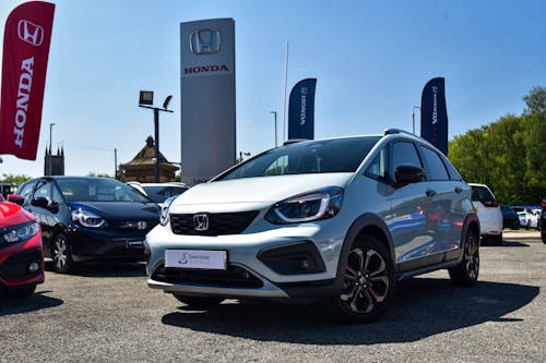 A silver Honda car is prominently displayed in an outdoor dealership lot, surrounded by other vehicles. Large banners and signs with the Honda logo are visible, with a clear blue sky in the background. The lot surface appears to be gravel or asphalt, and there is visible greenery on the side.