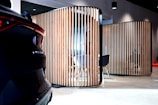 A modern interior space features wooden vertical slats forming partitioned areas, with a partially visible car on the left. The setting has contemporary furniture, including chairs and tables, visible through the slats. The lighting is soft, creating a sophisticated ambiance.
