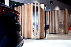 A modern interior space features wooden vertical slats forming partitioned areas, with a partially visible car on the left. The setting has contemporary furniture, including chairs and tables, visible through the slats. The lighting is soft, creating a sophisticated ambiance.