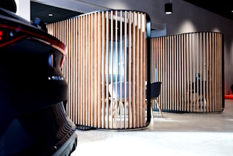 A modern interior space features wooden vertical slats forming partitioned areas, with a partially visible car on the left. The setting has contemporary furniture, including chairs and tables, visible through the slats. The lighting is soft, creating a sophisticated ambiance.