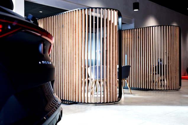 A modern interior space features wooden vertical slats forming partitioned areas, with a partially visible car on the left. The setting has contemporary furniture, including chairs and tables, visible through the slats. The lighting is soft, creating a sophisticated ambiance.