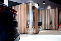 A modern interior space features wooden vertical slats forming partitioned areas, with a partially visible car on the left. The setting has contemporary furniture, including chairs and tables, visible through the slats. The lighting is soft, creating a sophisticated ambiance.