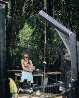 A woman wearing sunglasses holds a cat in her arms, standing in a shaded outdoor area surrounded by trees and various other cats. The environment includes netting and a table, suggesting a natural and serene setting.