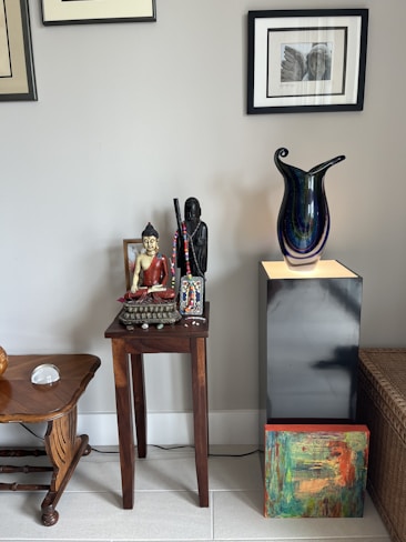 A wooden side table holds a Buddha statue and a figurine in dark colors, with intricate beading on display. Nearby, a large, colorful glass vase with a swirling pattern sits on a black pedestal. Framed artworks are mounted on a light gray wall, and a vibrant abstract painting rests against the pedestal. A wooden stool with curved legs and a decorative item beside it completes the arrangement.