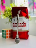 A package of yerba mate with a bright red and white design is on a table. In front of the package, there is a metal straw with a strainer at one end, known as a bombilla. Next to it, a solved Rubik's cube is visible. In the background, blurred green plants can be seen.