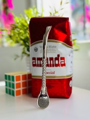 A package of yerba mate with a bright red and white design is on a table. In front of the package, there is a metal straw with a strainer at one end, known as a bombilla. Next to it, a solved Rubik's cube is visible. In the background, blurred green plants can be seen.
