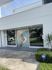 A modern house entrance with a decorative geometric metallic door featuring blue and beige accents. The entryway is flanked by large glass windows, reflecting nearby greenery and a parked white truck in the driveway. A small light fixture is mounted beside one of the windows. A neatly trimmed bush with an ADT security sign is positioned on the right, adjacent to a small palm plant. The surrounding area is paved with light stone tiles, and a well-maintained lawn is visible.