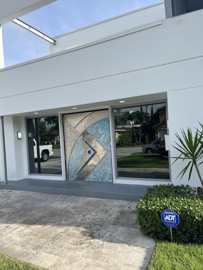 A modern house entrance with a decorative geometric metallic door featuring blue and beige accents. The entryway is flanked by large glass windows, reflecting nearby greenery and a parked white truck in the driveway. A small light fixture is mounted beside one of the windows. A neatly trimmed bush with an ADT security sign is positioned on the right, adjacent to a small palm plant. The surrounding area is paved with light stone tiles, and a well-maintained lawn is visible.