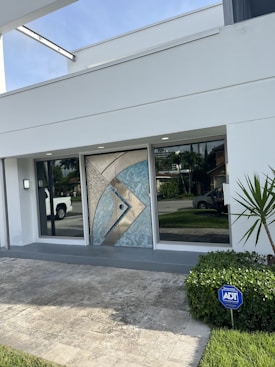 A modern house entrance with a decorative geometric metallic door featuring blue and beige accents. The entryway is flanked by large glass windows, reflecting nearby greenery and a parked white truck in the driveway. A small light fixture is mounted beside one of the windows. A neatly trimmed bush with an ADT security sign is positioned on the right, adjacent to a small palm plant. The surrounding area is paved with light stone tiles, and a well-maintained lawn is visible.