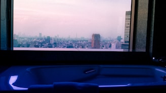 A modern bathtub is positioned in front of a large window offering a panoramic view of a cityscape. The skyline includes various buildings under a hazy sky during twilight, casting a serene ambiance.