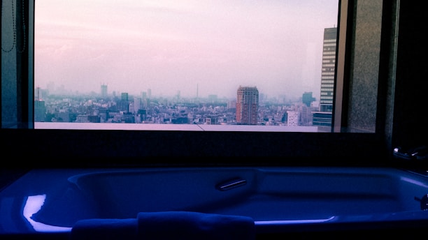A modern bathtub is positioned in front of a large window offering a panoramic view of a cityscape. The skyline includes various buildings under a hazy sky during twilight, casting a serene ambiance.