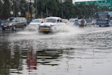 Several vehicles drive through a flooded street. The road is covered with water, creating splashes as cars, a taxi, a bus, and motorcycles navigate through. Adjacent palm trees and road signs are visible in the background.