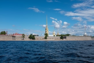 A fortress with a tall golden spire and domes is seen across a body of water. The structure is backed by a blue sky with scattered clouds. The foreground is filled with dark blue waves, while trees and a small sandy area line the base of the fortress walls.