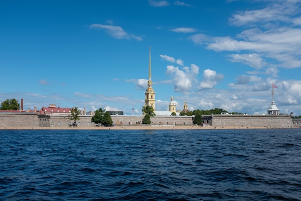 A fortress with a tall golden spire and domes is seen across a body of water. The structure is backed by a blue sky with scattered clouds. The foreground is filled with dark blue waves, while trees and a small sandy area line the base of the fortress walls.